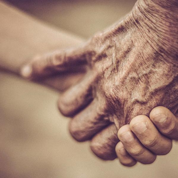 A close up of two hands joined against a blurred out, grey-brown background. The hands are wrinkled and aging.