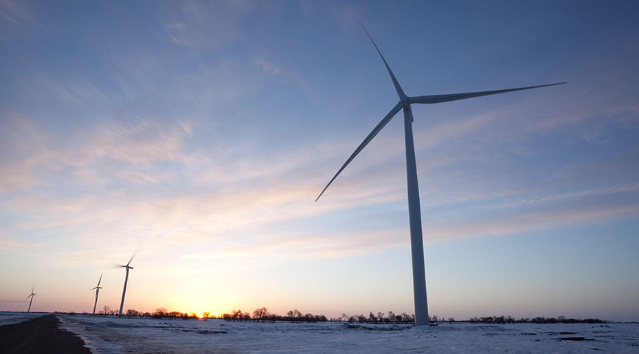 A low-angle perspective view of a wind turbine over a setting sun.