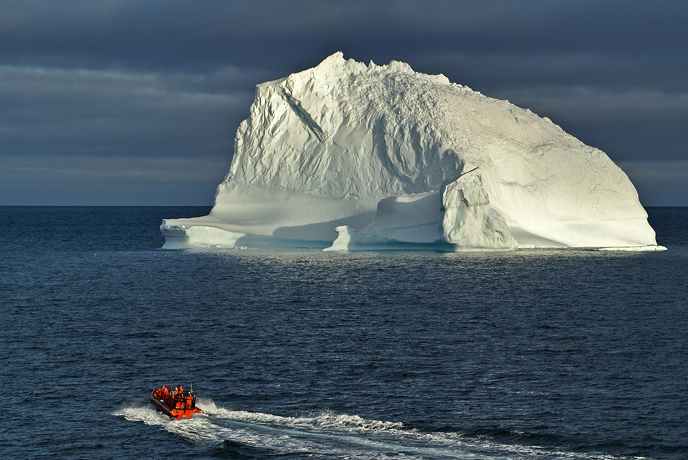 A landscape view of a small red boat in front of a massive iceberg.