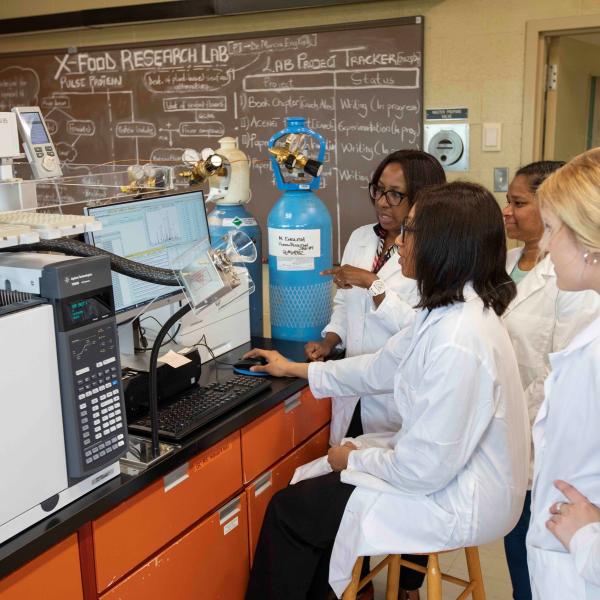 Four people gather around a mass spectrometer on a lab desk.