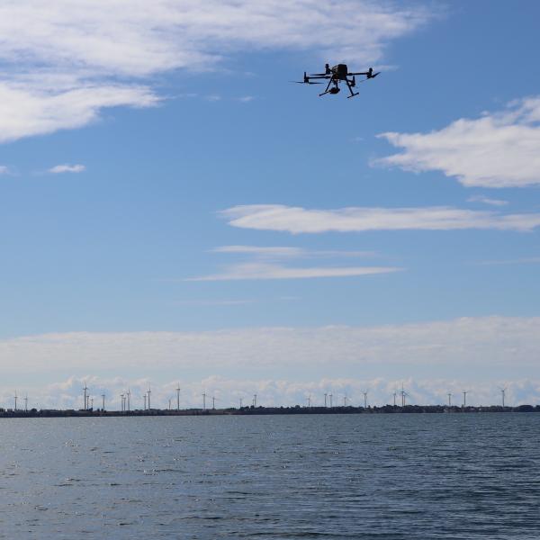 A quadrotor UAV flies in the upper right of the photo; in the water below floats a small uncrewed vessel Greef and her team plan to use as a recharging station.