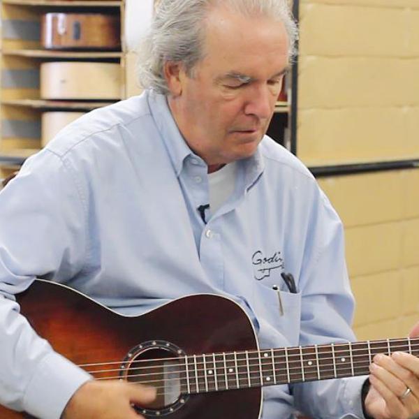 Robert Godin sits inside a bright warehouse, playing one of the guitars made by his company, Godin Guitars. The shelves behind him are full of guitars.