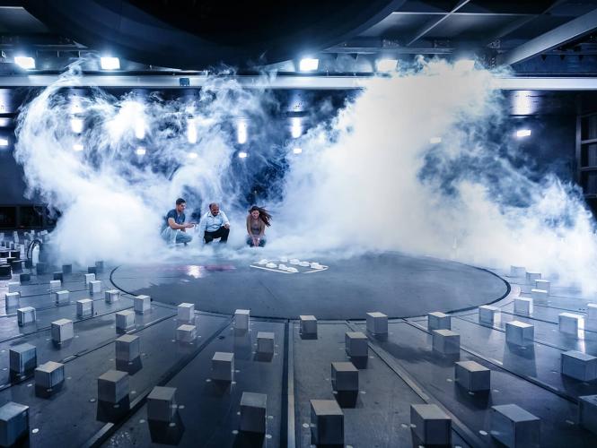 Three people sitting in a test chamber in front of a miniature town, surrounded by gusts of white smoke and wind.