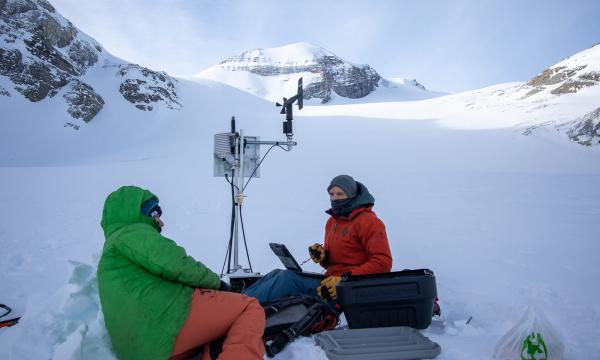 Two people in winter clothing sit in the snow in the mountains with research equipment around them.