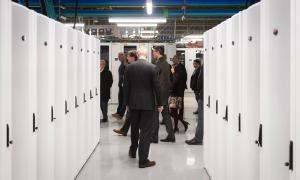A group of people in business attire walk among a series of tall, white, metal cabinets. CAPTION: The cylindrical structure that now houses Université Laval’s Colosse supercomputer was first built in 1962 as a particle accelerator, which was dismantled in 2006. CREDIT: Wikimedia Commons ALT TEXT: A cylindrical concrete building covered in a metal cage and green ivy.