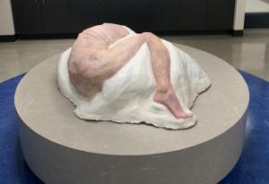 A sculpture of a human body on its side, curled into a ball lies on a circular dais.