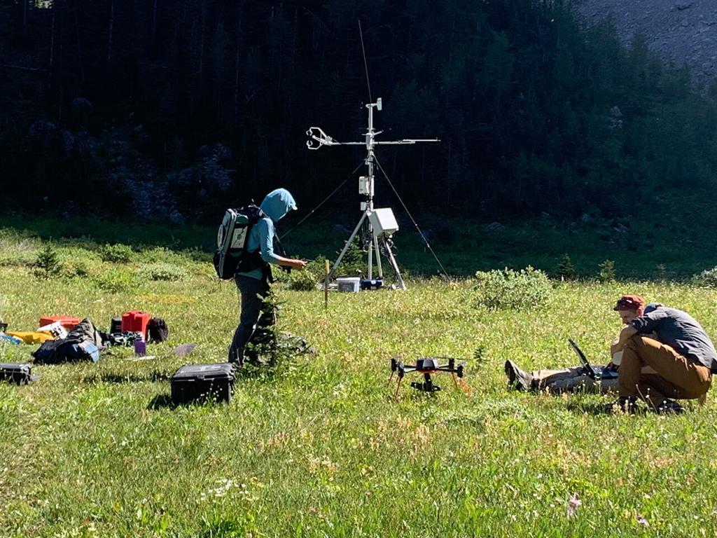 Two people, one standing, one seated in a grassy area with pieces of research equipment around them.