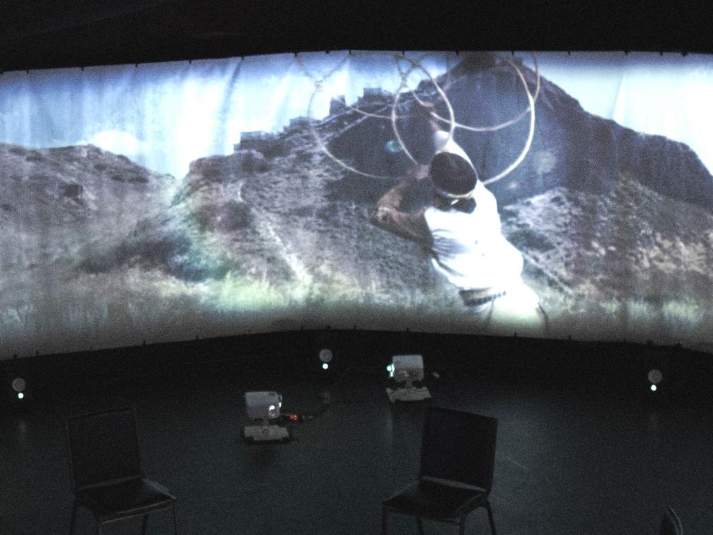 A person hoop dances on a panoramic screen in a large circular space.