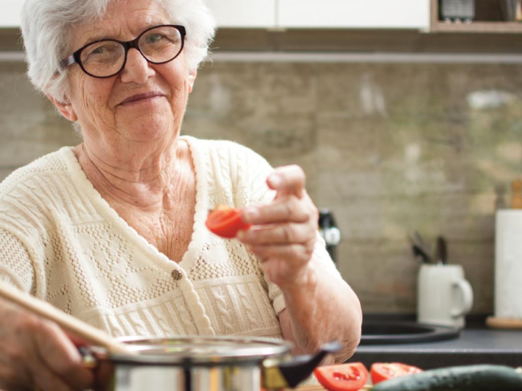 Un quartier de tomate à la main, une femme aux cheveux blancs se tient debout devant un chaudron sur une cuisinière