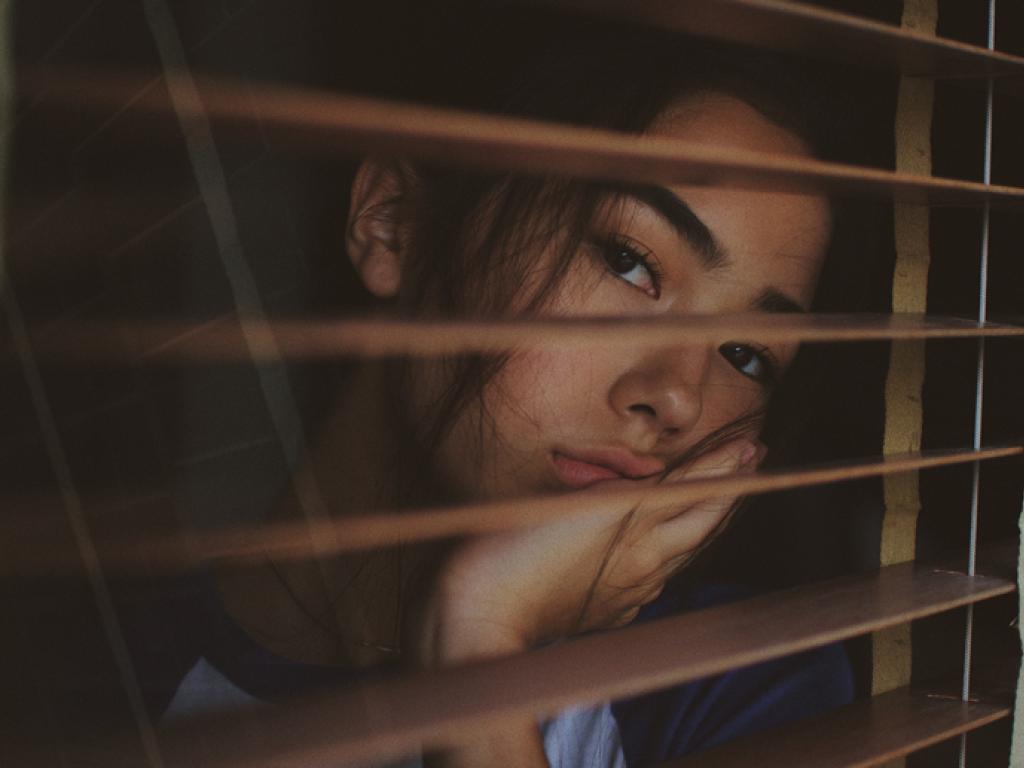 Une jeune femme regarde par la fenêtre à travers les volets ouverts, l’air ennuyé