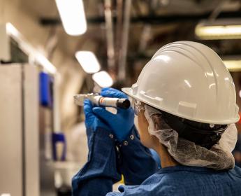 Une personne debout dans un laboratoire, portant un casque de sécurité et des gants bleus, regarde dans un appareil cylindrique de petite taille