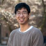 A person wearing a grey sweater posing for a photo, in front of a blurry floral background.